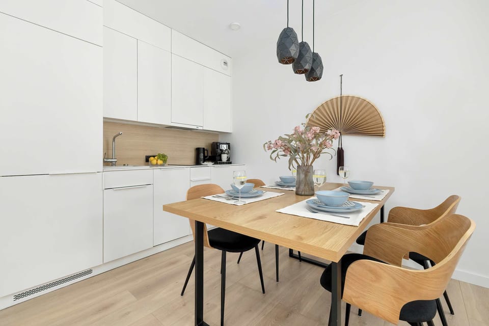 A wooden dining table paired with four warm brown chairs sits in front of a white kitchen wall. A stylish woven pendant lamp hangs above the table.