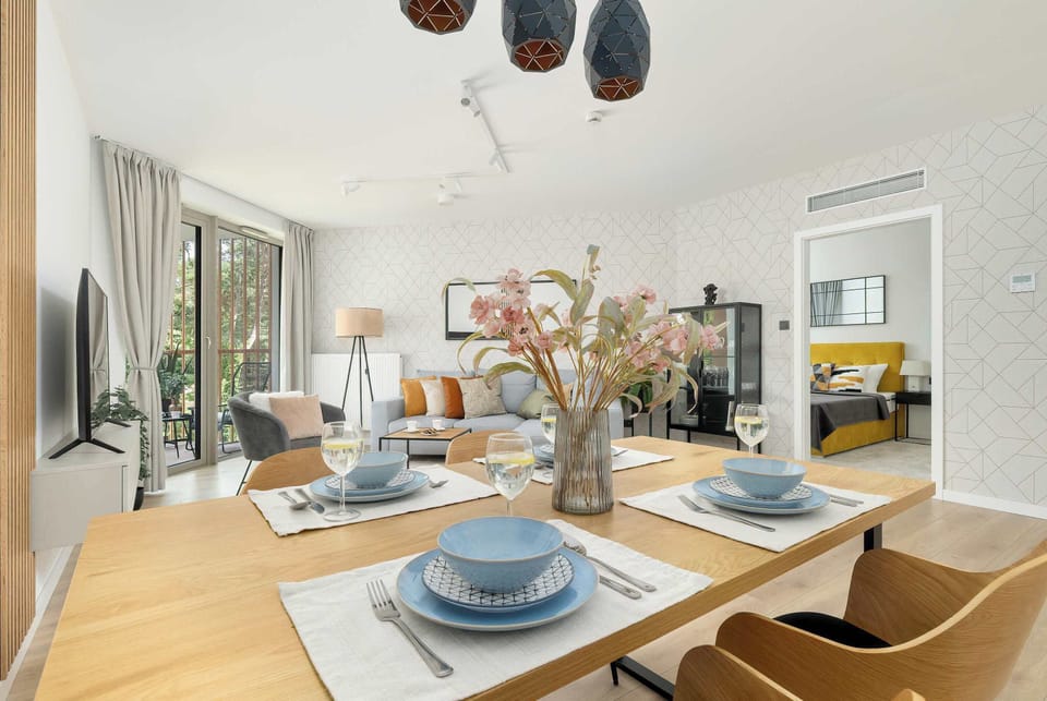 A wooden dining table set for four features light blue place settings and a vase of flowers in the center. The open doorway reveals the bedroom.