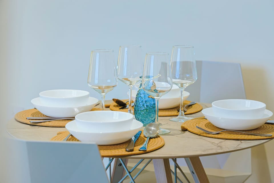 Your dining table awaits, beautifully set with crisp white bowls, sparkling wine glasses, and natural woven placemats—perfect for enjoying leisurely meals together in coastal style.