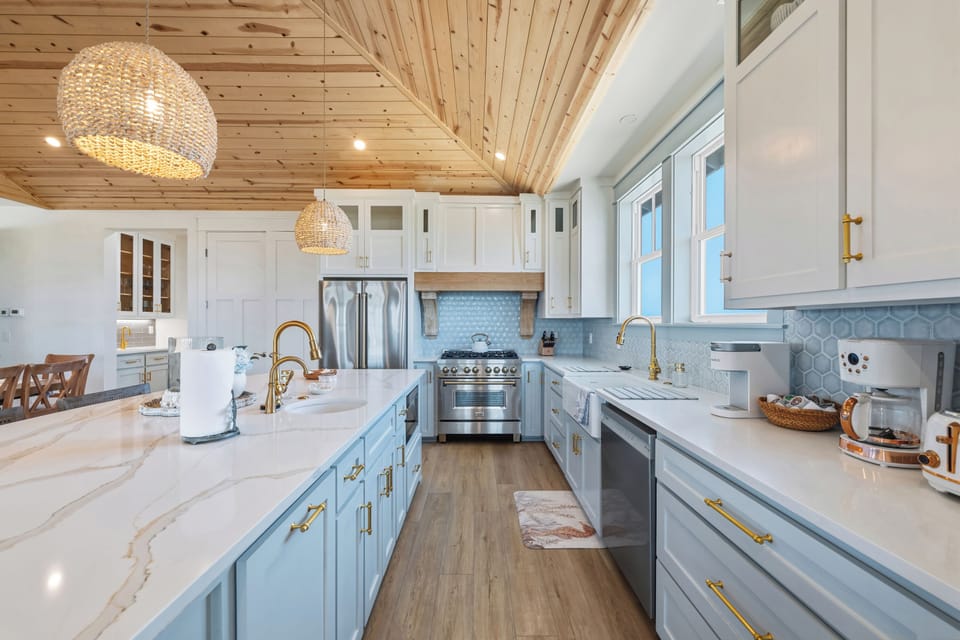 A spacious chef’s kitchen featuring elegant countertops, a natural quartz island, soft blue cabinetry with brushed gold hardware and fixtures, stainless steel appliances, and a vaulted wood-plank ceiling that adds warmth and coastal character.