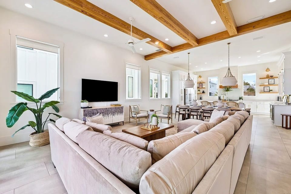 Sunlit 2nd-floor Great Room with plush sectional seating, 75" 4K Smart TV, exposed wood beams, and open chef’s kitchen. Open to balcony dining and lounging.