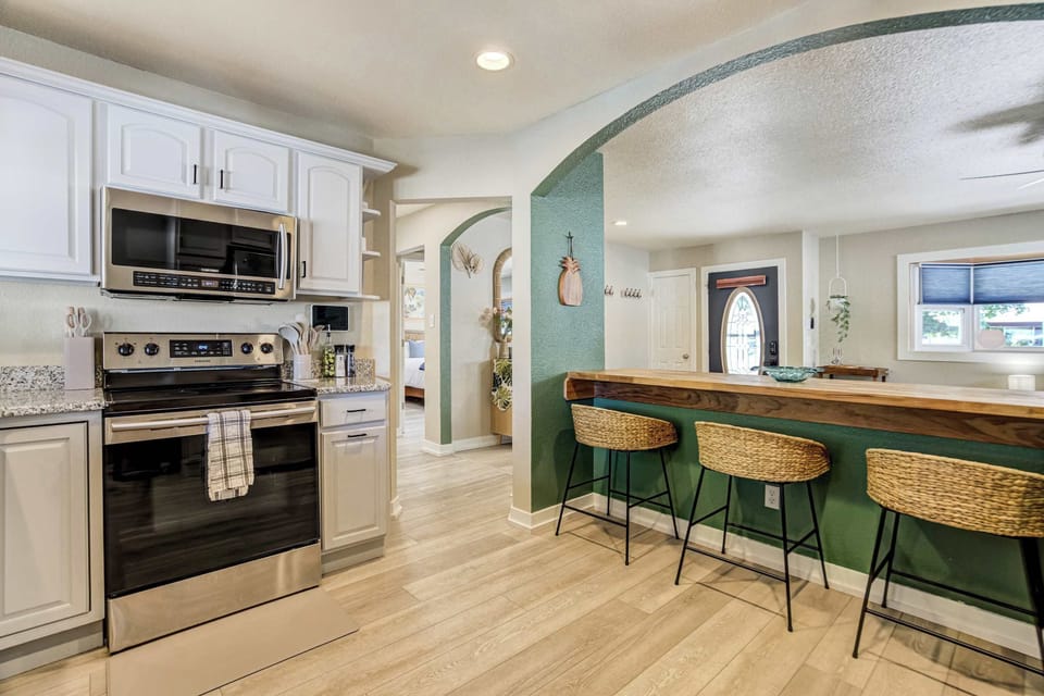 The kitchen sits open to the living room, so whoever is cooking stays part of the group. Stainless appliances, granite counters, and white shaker cabinetry line the wall, with the arch framing the bar and seating beyond.