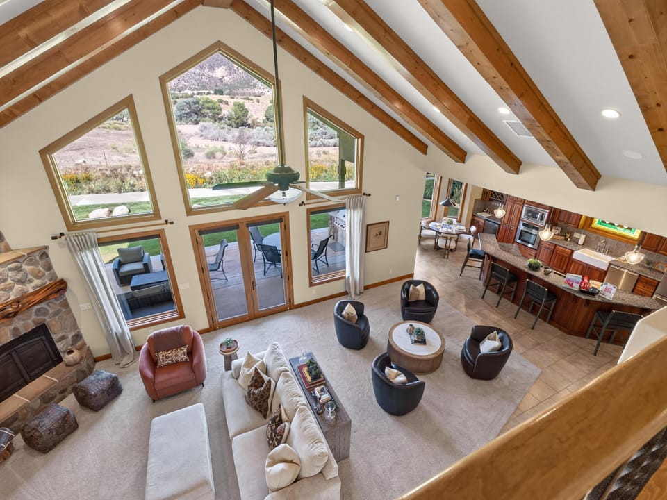Living room with vaulted wood beams, stone fireplace, and large triangular windows overlooking the patio and landscape.