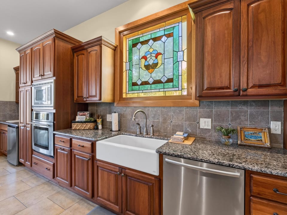 Farmhouse sink sits below a stained glass window, surrounded by granite countertops and wood cabinetry.