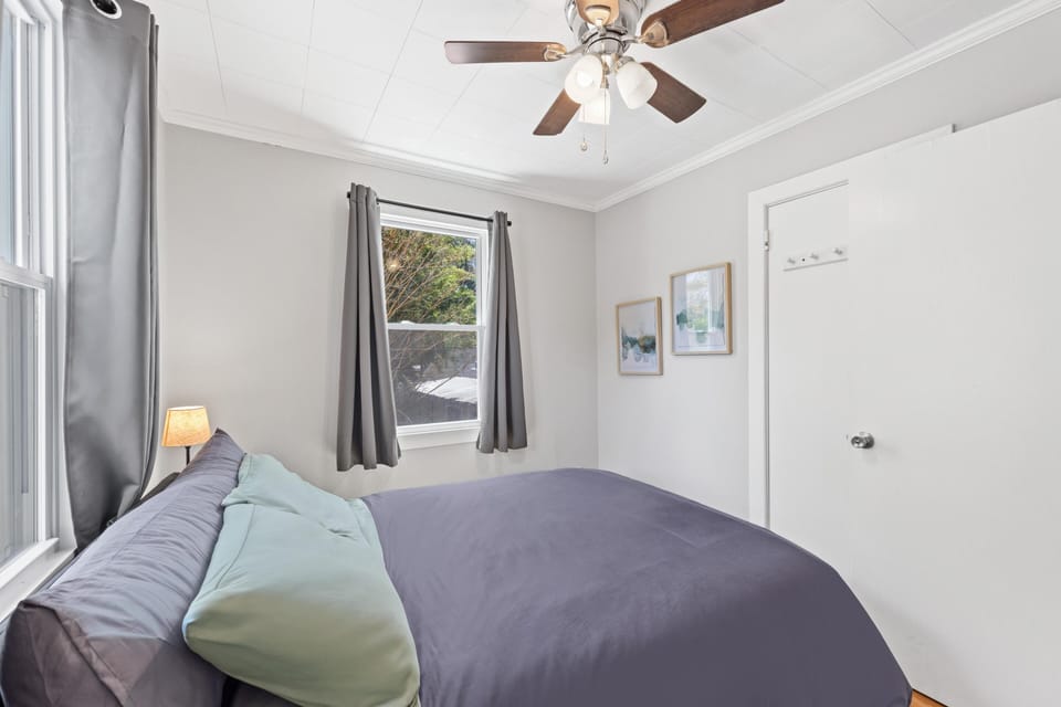 Bedroom with gray curtains framing two windows and a ceiling fan with wood-toned blades overhead.