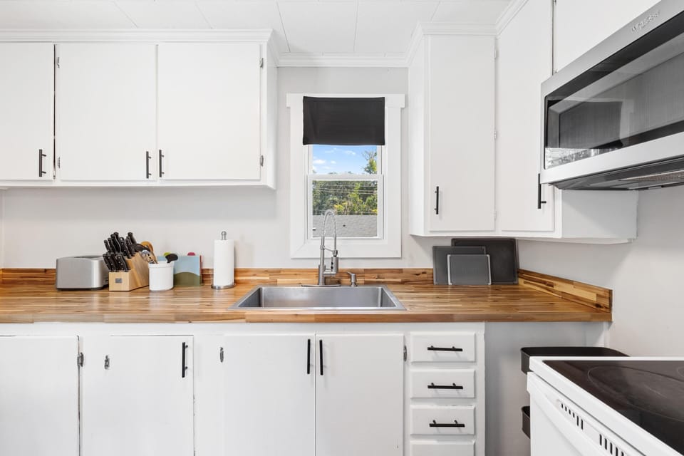Kitchen countertop features natural wood grain with stainless steel sink beneath a window with black shade.