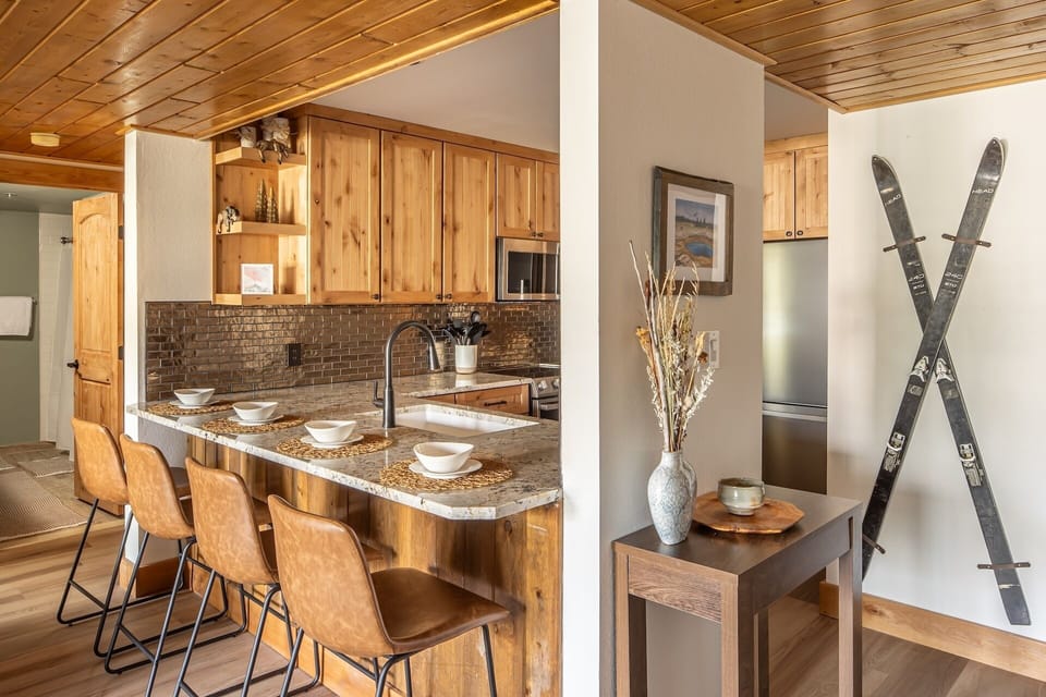 Your alpine retreat awaits with a warm wooden kitchen featuring a granite breakfast bar where you can gather for morning coffee before hitting the slopes—vintage skis on the wall set the cozy mountain mood.