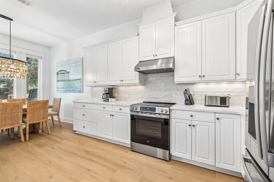 Step into your bright, coastal-inspired kitchen with gleaming stainless appliances, white cabinetry, and warm wood floors. Your morning coffee brews beside a dining nook where you'll gather around the table, planning adventures ahead.