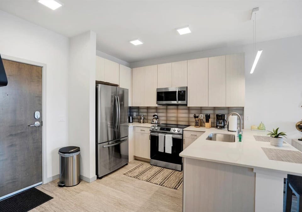 Modern kitchen with stainless steel appliances, sleek cabinetry, and a spacious island featuring a deep sink and breakfast bar. Warm wood accents and stylish lighting create a clean, contemporary cooking and dining space.