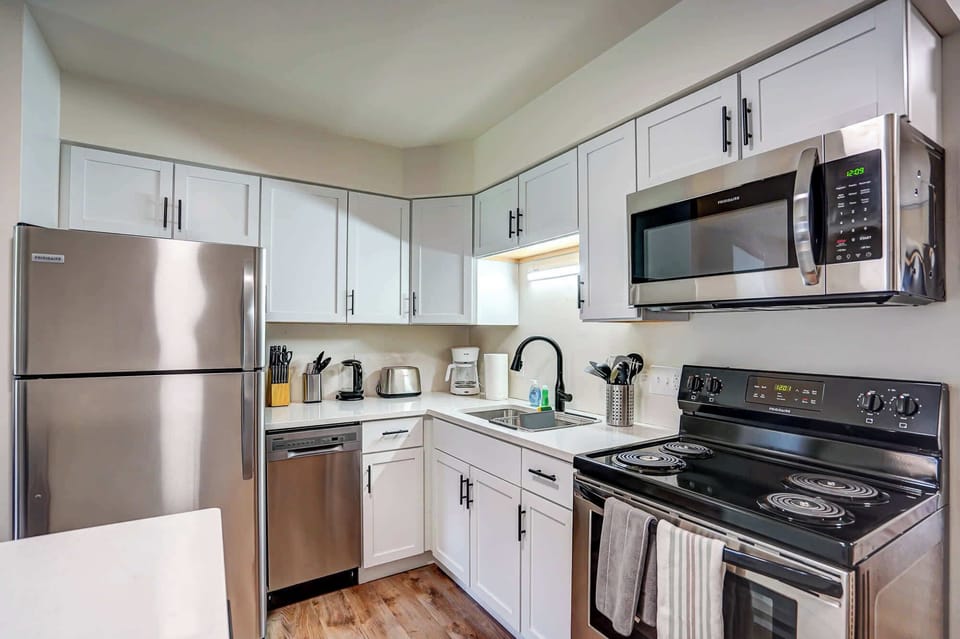 Modern kitchen featuring stainless steel appliances, sleek white cabinetry, and wood-style flooring. Fully equipped with essentials like a coffee maker, toaster, and cookware—perfect for preparing home-cooked meals during your stay.