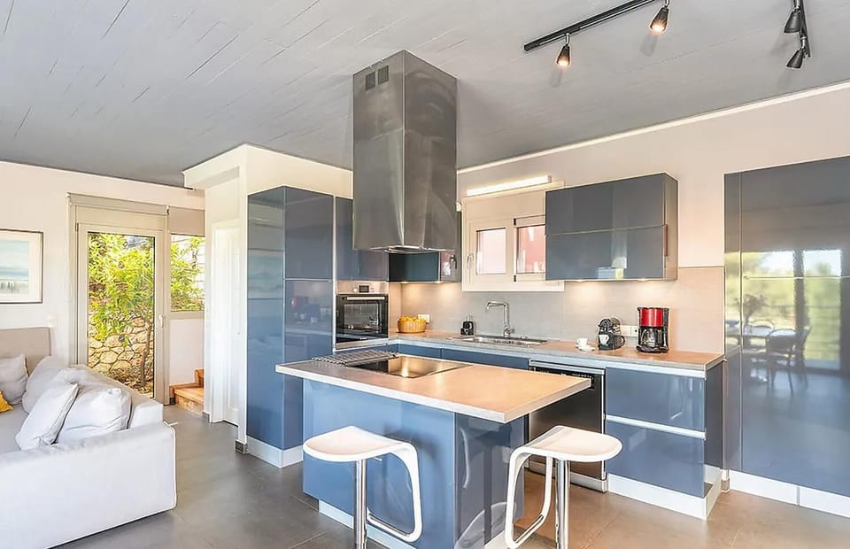 A sleek contemporary kitchen features glossy blue-gray cabinets, a central island with white bar stools, and a large stainless steel range hood under a muted gray ceiling.