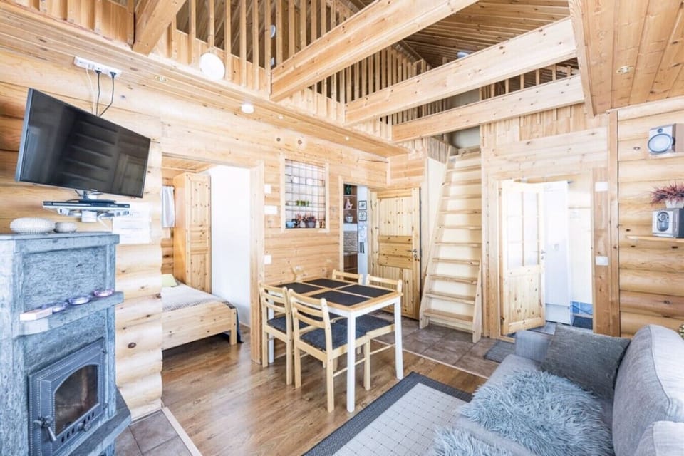 Bright open-plan living space with a stone fireplace, wall-mounted TV, dining table and stairs up to the loft.