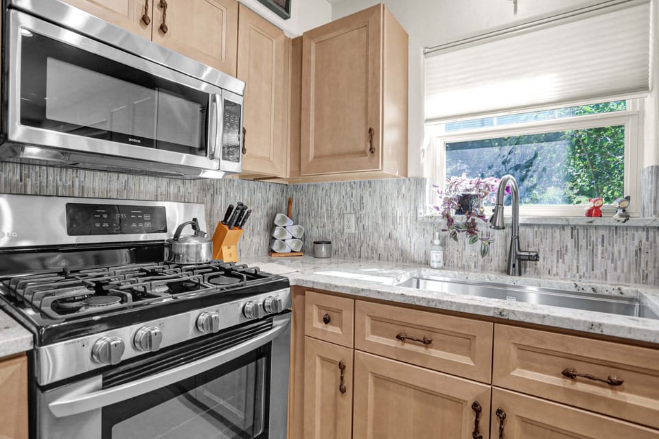 A well-lit, contemporary kitchen showcases light wood cabinetry, a mosaic tile backsplash, and stainless steel appliances including a gas range and an over-the-range microwave.