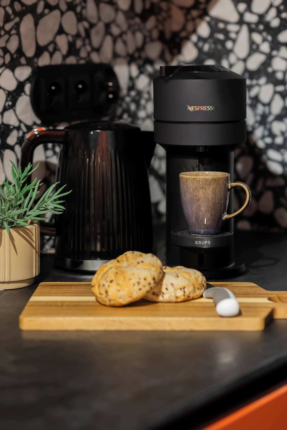 A close-up shot focuses on a black coffee maker pouring an espresso beside fresh baked pastries on a wooden serving board.