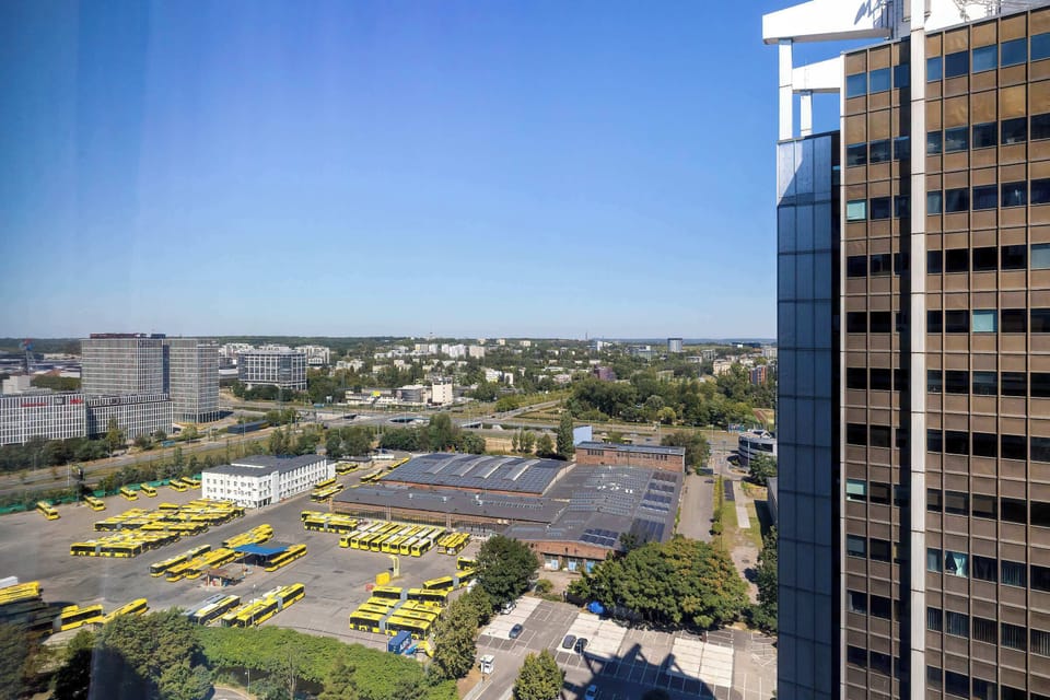 A high-angle view from an upper floor looks down on an urban cityscape, neighboring commercial rooftops, and a blue horizon.