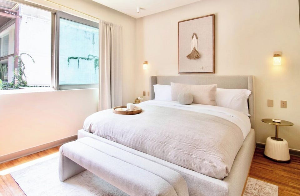 Natural light enters through the generous window, while the upholstered bench at the foot of the bed offers a comfortable place to sit and get ready. A calm neutral palette keeps the room feeling bright and effortlessly sophisticated.