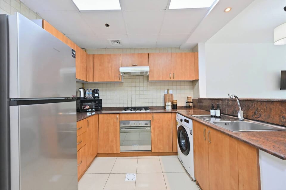 Compact, fully equipped kitchen with warm wooden cabinets, granite countertops, and tiled backsplash. Features a built-in oven, gas cooktop, microwave, and washing machine, offering a practical and functional space for everyday cooking.