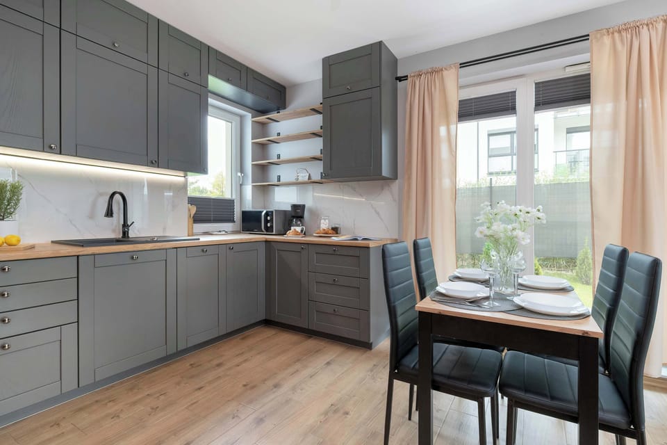 The open-concept kitchenette is equipped with matte grey cabinetry and built-in appliances. A four-person dining table with upholstered black chairs creates a dedicated dining corner. Warm wood flooring ties the kitchen seamlessly to the living area.