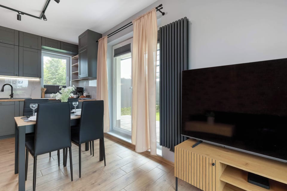 The dining and entertainment corner features a black dining set alongside a wooden TV console with a flat-screen television. Floor-to-ceiling sheer curtains frame the glass door to the private garden. The layout maximizes open-plan functionality.