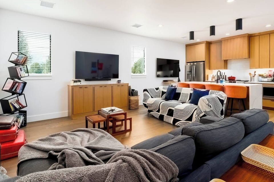 This modern open-concept space seamlessly blends living room and kitchen areas. Features include a gray sectional sofa, wall-mounted TV, unique zigzag bookshelf, and honey-toned wooden cabinetry with white countertops and stainless appliances.
