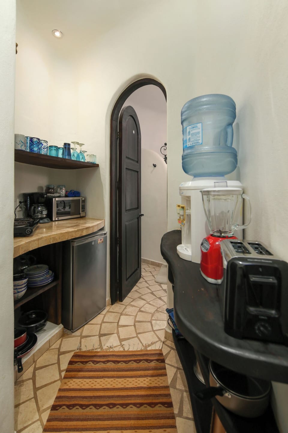 Kitchenette corridor with open shelves, blue glassware, coffee maker, microwave, mini fridge, water cooler, blender, and toaster