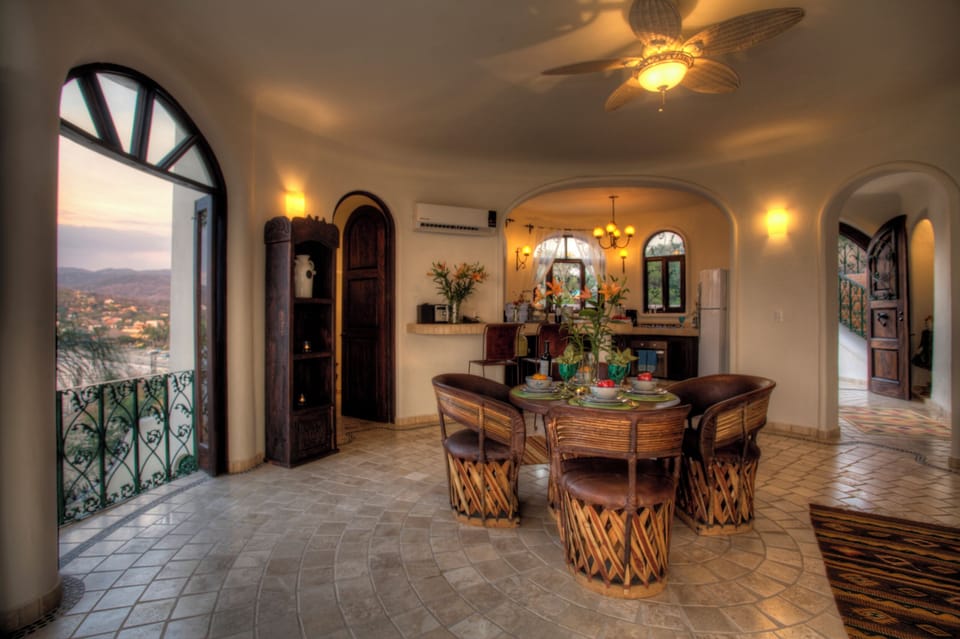 Open dining and kitchen with bamboo chairs, fresh lilies, and arched balcony door at dusk
