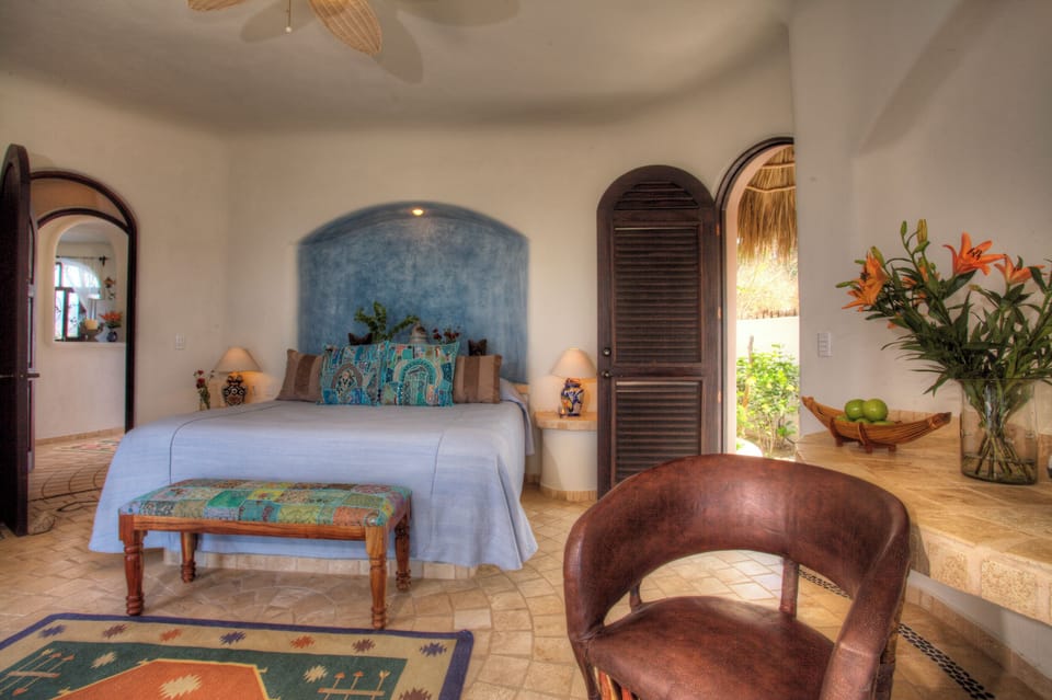 King bedroom with blue plaster headboard niche, patchwork bench, bamboo sitting chair, and patio door