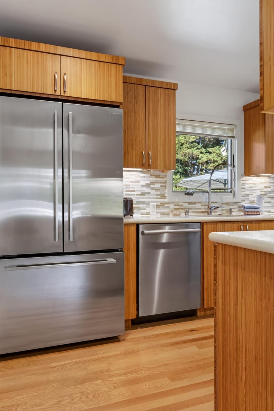 Under-cabinet lighting highlights the neutral mosaic backsplash above the sink, where tree-top views fill the window over the counter.
