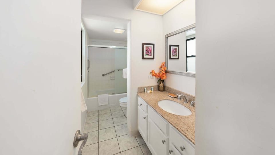 A bright guest bathroom featuring a single-sink vanity with granite countertop, large silver-framed mirror, decorative floral touches, and tiled flooring.