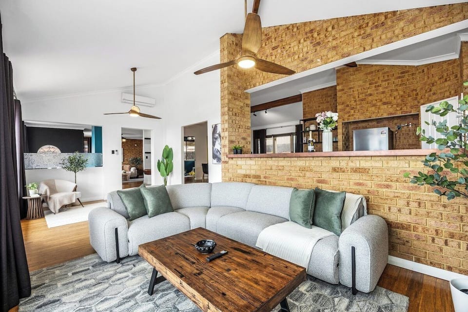 Unwind in this stylish, open-concept living area featuring a light gray curved sectional sofa, green accent pillows, a rustic wood coffee table, exposed brick walls, and vaulted ceilings with wooden ceiling fans.