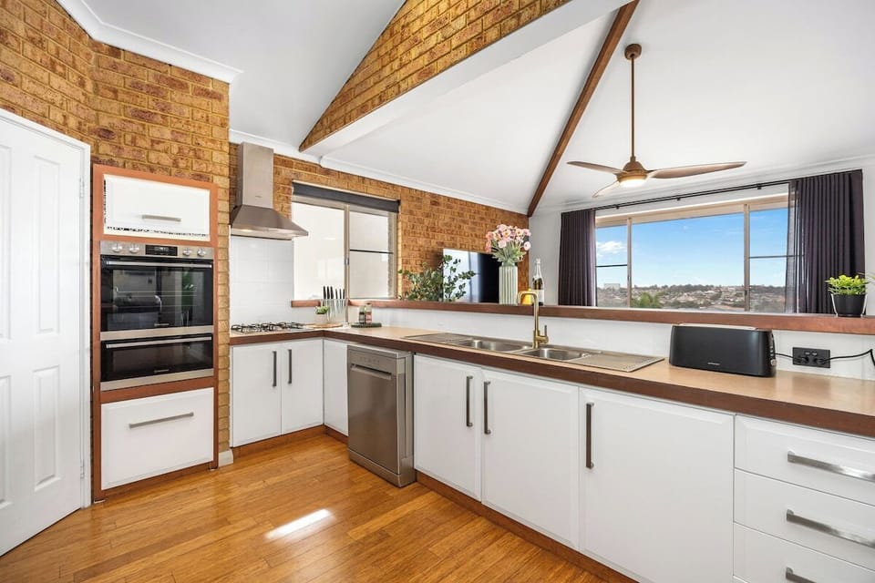 Prepare meals with ease in this well-appointed kitchen, featuring clean white cabinetry, warm wooden countertops, integrated double wall ovens, a stainless steel dishwasher, exposed brick walls, and vaulted ceilings with exposed beams.
