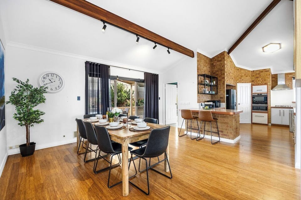Gather for meals in this open-concept dining and kitchen space, featuring a light wood dining table with modern black chairs, exposed brick accents, high vaulted ceilings with wooden beams, track lighting, and a breakfast bar with seating.