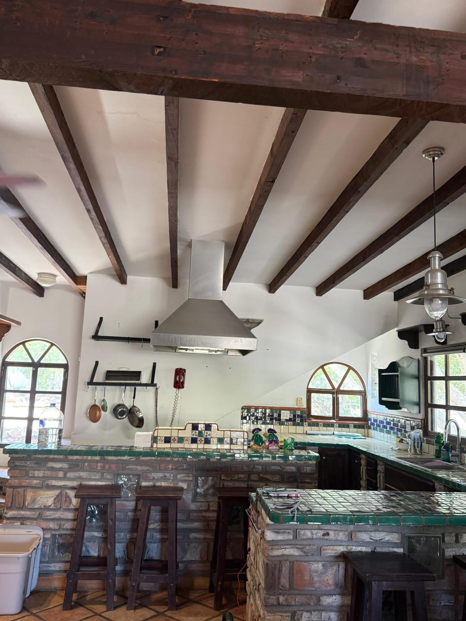 A focused interior view of the kitchen cooking zone, showcasing the built-in brick cooktop counter, large stainless steel range hood, and an overhead hanging rack for pots and pans.