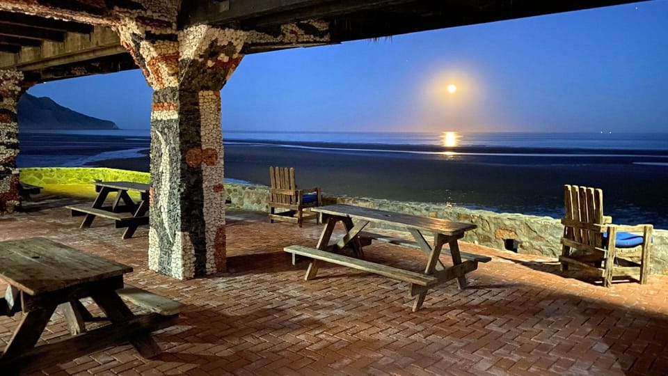 A tranquil sunset view from the villa's covered brick patio, highlighting rustic stone pillars and a wooden picnic bench looking out over a calm, moonlit ocean horizon.