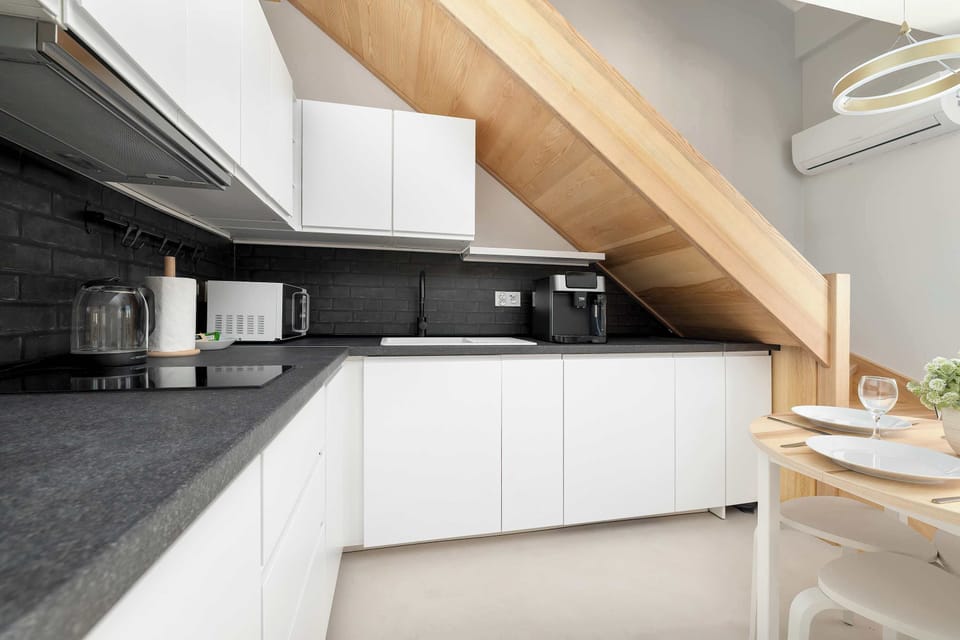 Clean white kitchen cabinetry fits neatly under the slant of a wooden staircase, maximizing the functional layout. The dark countertop provides plenty of preparation room for home cooking.