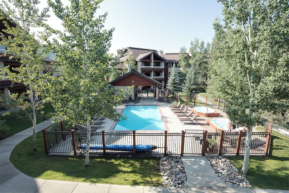 Multi-building vacation rental complex nestled among birch and evergreen trees, featuring an outdoor swimming pool, hot tub, and lounge deck surrounded by lush green lawns and natural landscaping.