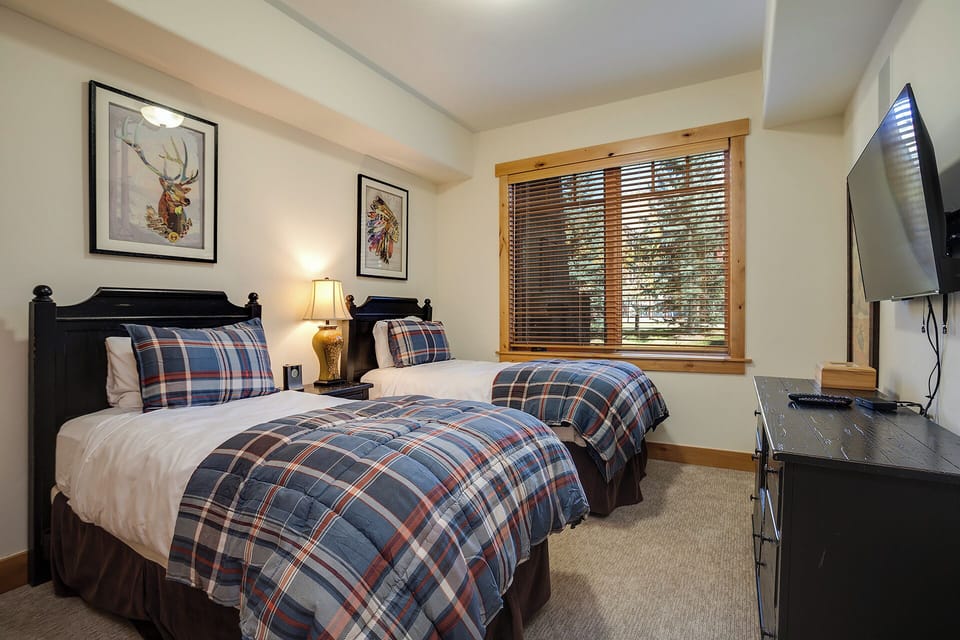 Sink into crisp white linens topped with plaid throws, where two comfortable beds await your arrival. Natural light pours through wooden blinds, framing peaceful forest views.