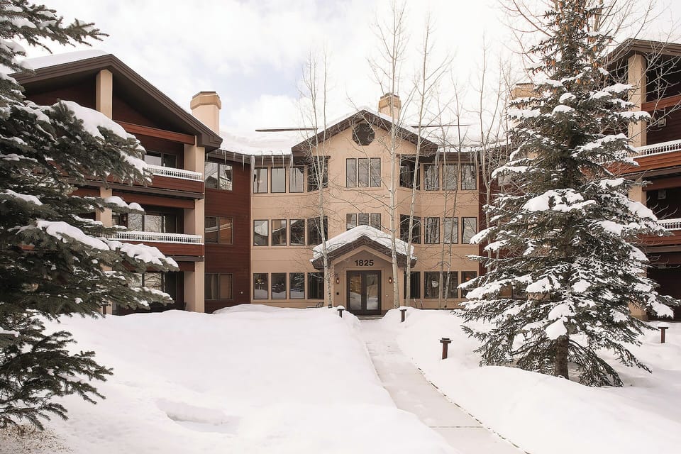 Nestled among snow-laden evergreens, the property's welcoming entrance at 1825 offers alpine charm and easy winter access. The multi-story building stands ready to host a cozy mountain retreat.