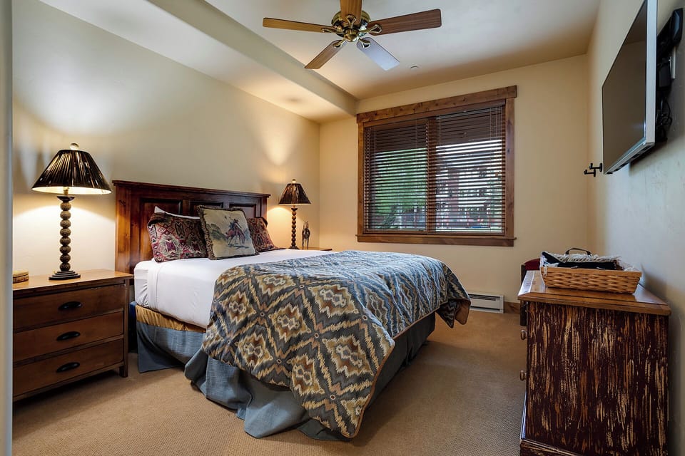 Sink into your cozy bed under vaulted ceilings with a ceiling fan and warm lighting. Natural light filters through wooden blinds, while rustic furnishings and a wall-mounted TV complete your restful retreat.