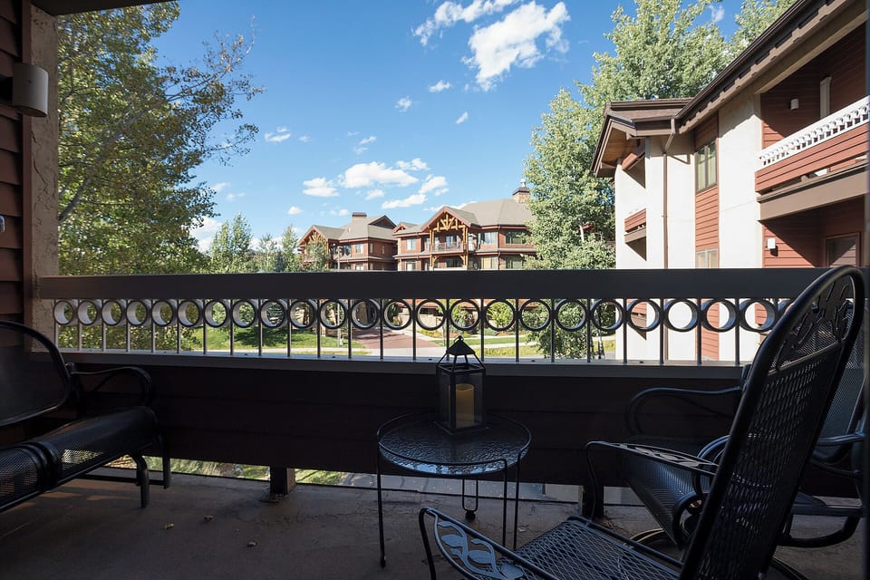The property is framed by trees and open skies, with neighboring buildings and a sunny blue sky visible from the terrace—ideal for relaxing outdoors during your stay.