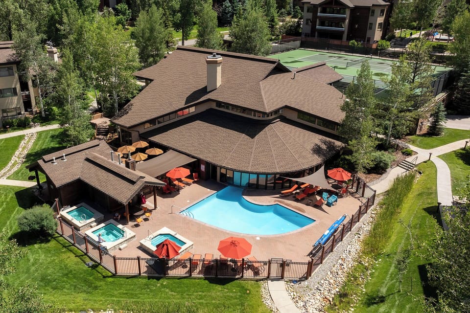 Aerial view of a mountain-side vacation property featuring a heated pool, hot tub, and tennis courts surrounded by mature pines and manicured lawns.
