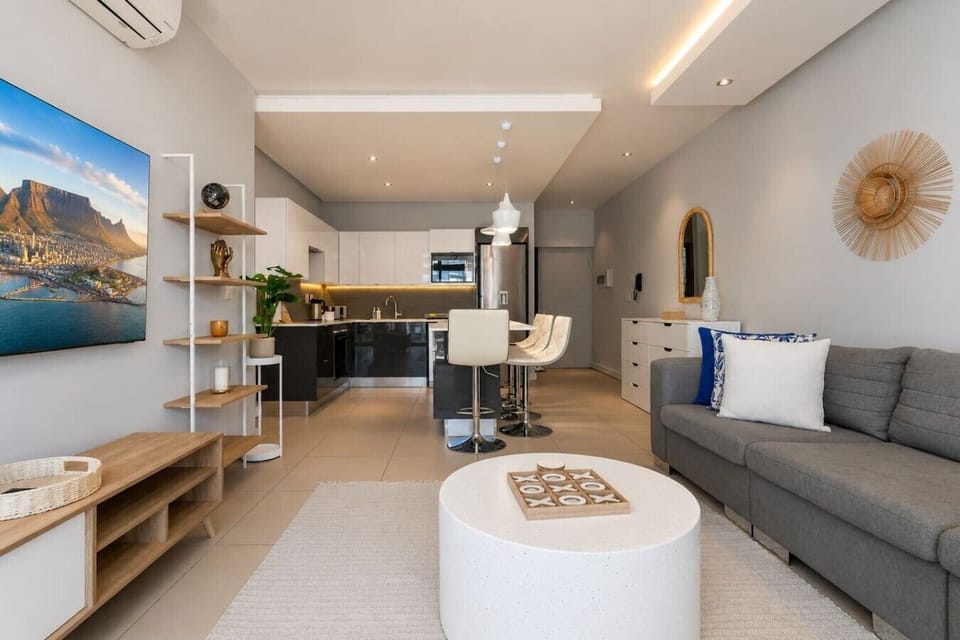 Living Room: Contemporary open-plan lounge with a comfortable grey sofa, round coffee table, wall-mounted TV, and curated display shelves. The seamless layout flows toward a stylish kitchen with an island and bar seating.