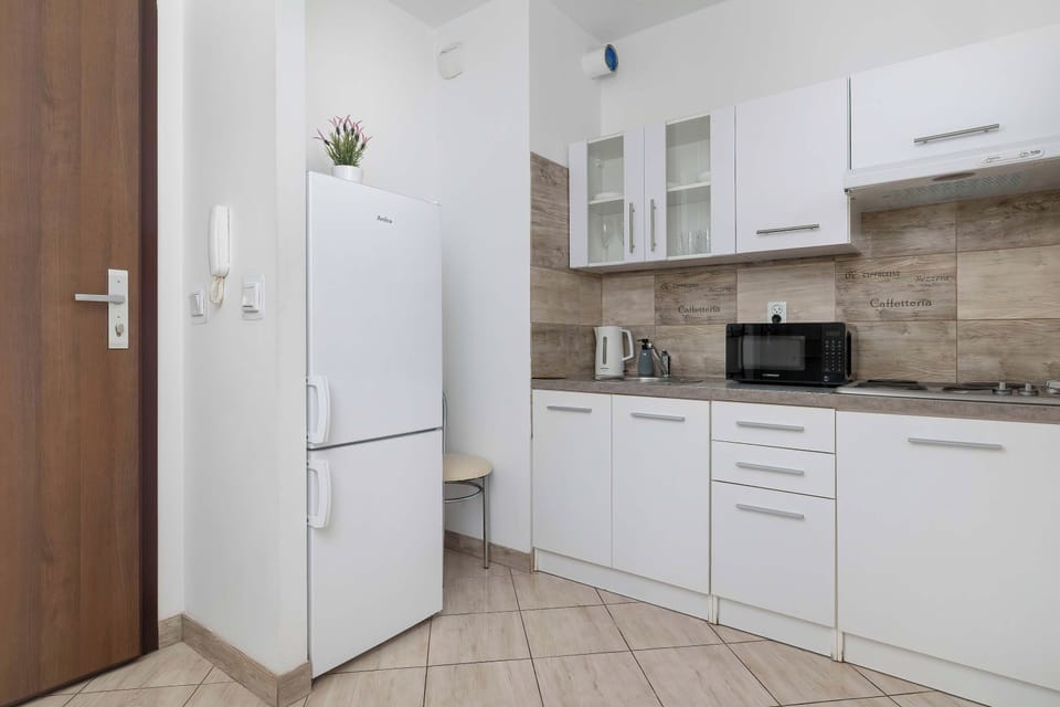 A bright kitchen corner features a white refrigerator, microwave, and cabinetry set against neutral beige wall tiles.