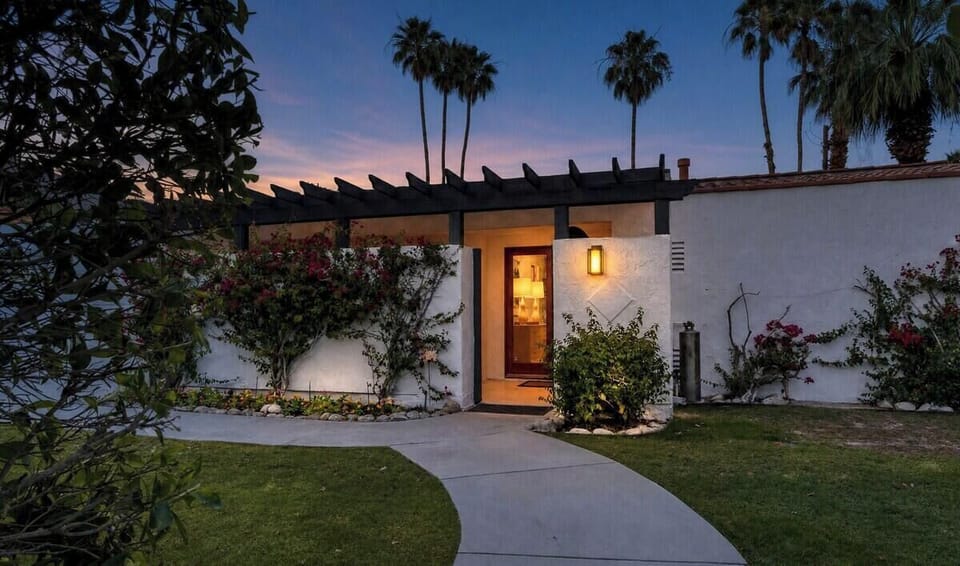 A welcoming desert retreat framed by swaying palms and lush landscaping, offering a tranquil arrival experience as twilight paints the sky in soft hues.