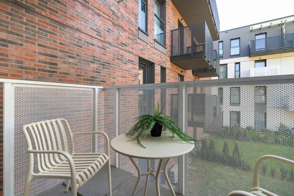 Another balcony perspective shows a small round table flanked by two outdoor chairs, providing a spot for outdoor relaxation. The building's exterior architecture features prominent sections of exposed red brickwork.