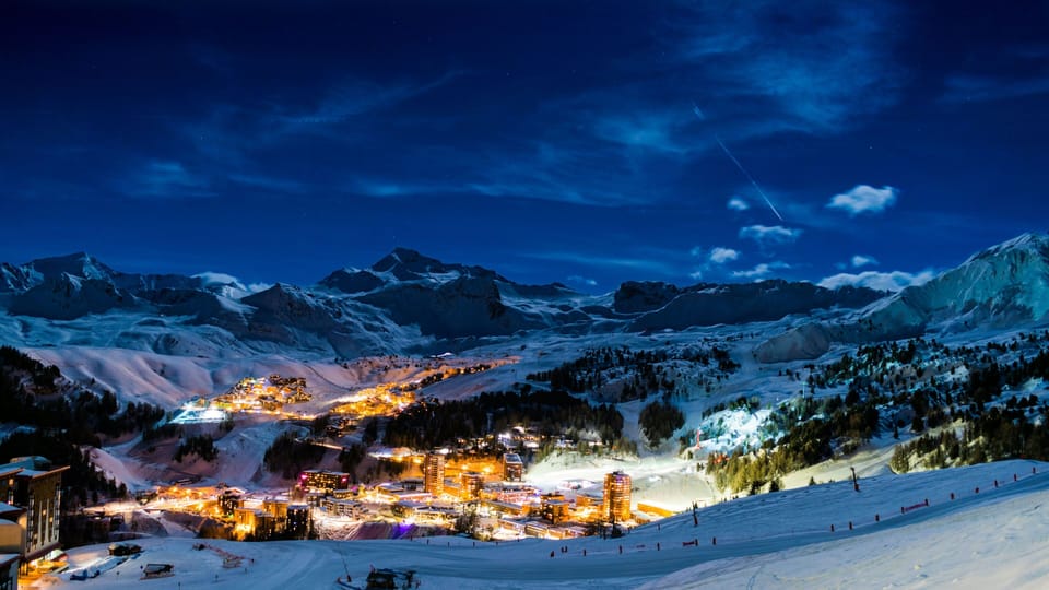 Aerial view of village illuminated at night against a backdrop of snow-covered mountain peaks
