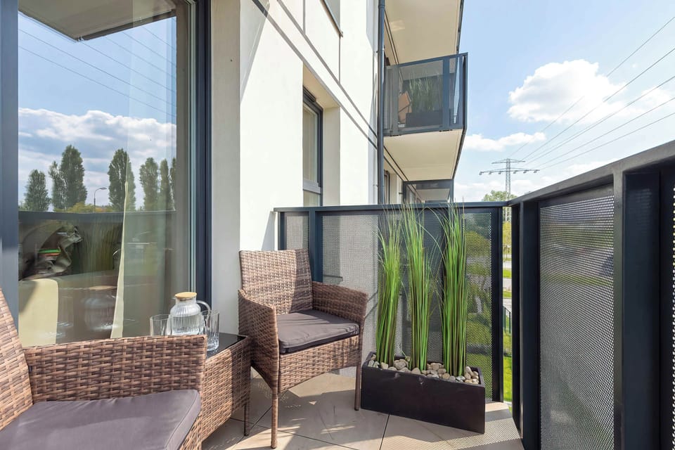 A balcony furnished with outdoor seating overlooking neighboring buildings and greenery. Glass panels and a covered structure provide a comfortable outdoor setting.