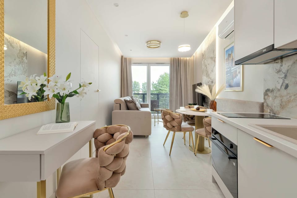 A spacious open-plan interior combining a beige sofa, dining area, and modern kitchen. Large windows and light furnishings give the room an airy, elegant feel.