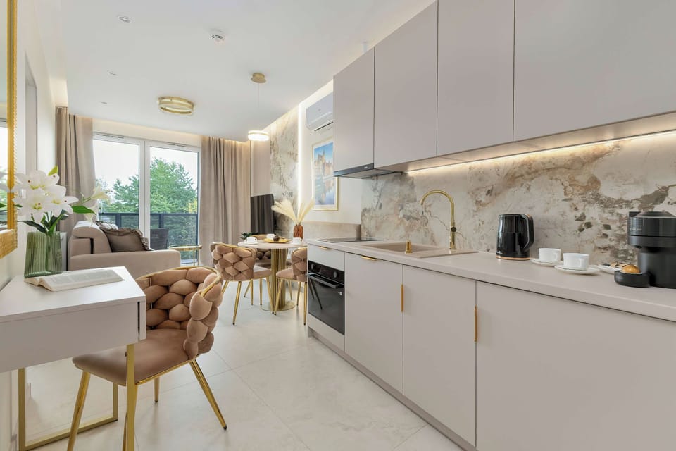 A bright open-plan kitchen with white cabinetry, a marble-effect backsplash, and a dining area with upholstered chairs. Large windows bring natural light into the space.