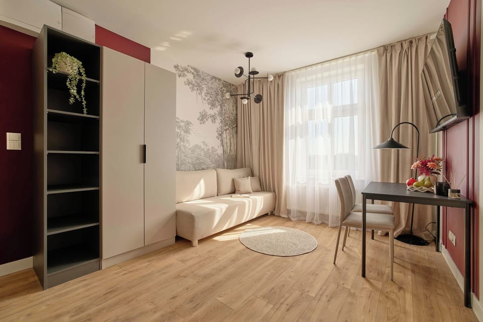 Stylowy i komfortowy salon z wygodną sofą, dużymi oknami i elegancką strefą wypoczynku. Ciepła drewniana podłoga, dekoracyjna tapeta, burgundowe akcenty i nastrojowe oświetlenie nadają wnętrzu charakter.